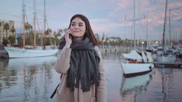Smiling and cheerful beautiful young brunette girl receives a call on her smart phone and talking. She stays alone on peaceful romantic backround of sea port with parked boats at early morning
