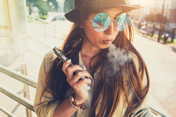 Pretty young hipster woman in black hat vape ecig, vaping device at the sunset. Toned image.