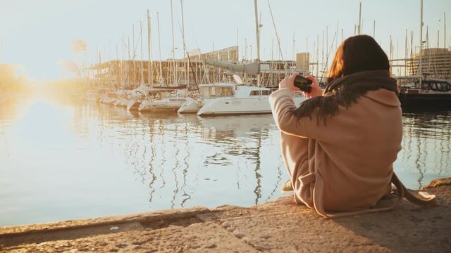 Backview on girl in warm coat sitting alone in port and taking picture of sunrise in front of parked yachts in small marina at early autumn morning.