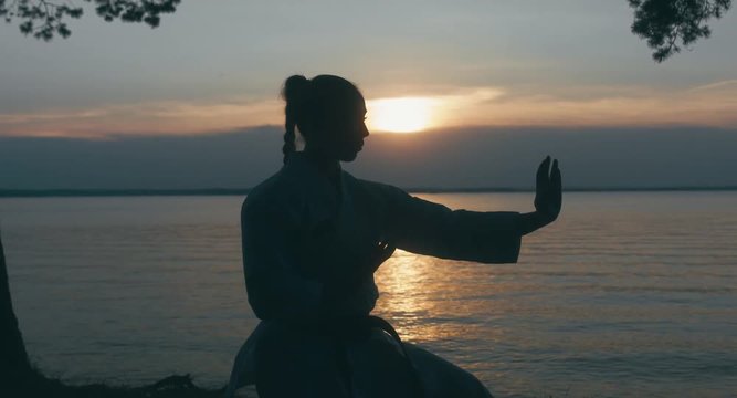 MED Caucasian professional female athlete wearing kimono practicing karate near large lake, sunset shot. 4K UHD, 60 FPS SLO MO