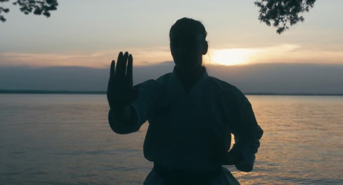 CU Caucasian professional female athlete wearing kimono practicing karate near large lake, sunset shot. 4K UHD, 60 FPS SLO MO