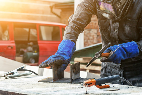Man welder in a welding mask, building uniform and blue protective gloves brews metal welding machine on the street construction, in the background an old brick building, red old van, the sun shines