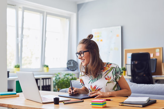 Cheerful attractive young female graphic designer smiling and working at her desk in modern office
