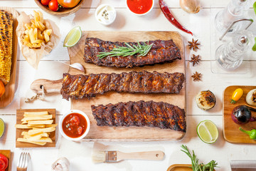 Spicy hot grilled spare ribs from a summer BBQ served with chips, corn and fresh tomatoes on an old wooden cutting board, top view