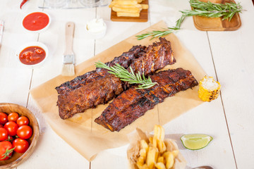 Spicy hot grilled spare ribs from a summer BBQ served with chips, corn and fresh tomatoes on an old wooden cutting board