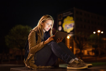 Woman in the city at night holding tablet, reading something.