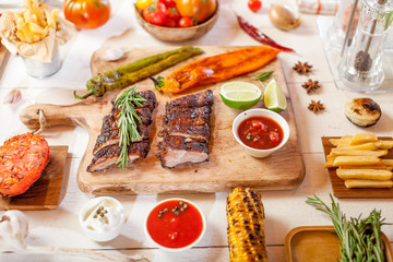 Spicy hot grilled spare ribs from a summer BBQ served with chips, corn and fresh tomatoes on an old wooden cutting board