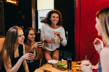Four cheerful girlfriends having fun chatting and laughing eating and drinking in fast food restaurant
