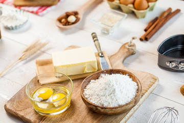 Baking ingredients in rural kitchen - dough recipe ingredients (eggs, flour, milk, butter, sugar) and rolling pin on wooden white table.