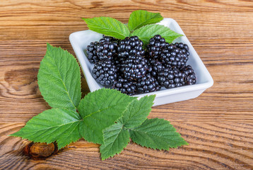Sweet juicy blackberries. Decorative bramble leaves. Rubus. Delicious black forest berries in white square porcelain bowl on a wood background. Green natural twigs. Healthy organic vegetarian dessert.