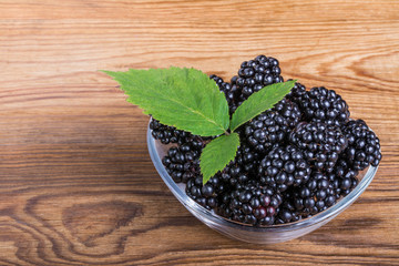 Heap of blackberries in glass bowl. Fresh juicy fruit with a decorative green leaf on a wooden background.