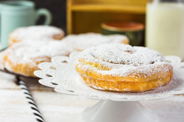 Traditional Spanish or Philippine pastry ensaimada. Powdered, on cooling rack and white cake stand. Bottle of milk, straw. Cozy rustic kitchen interior.