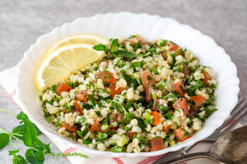 Tabbouleh salad with bulgur, tomatoes, parsley, green onion and mint in plate on grey stone table.