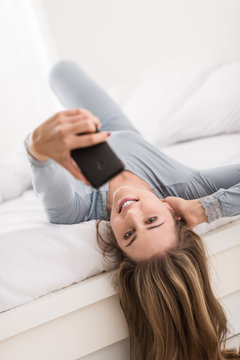 Young smiling woman lying in bed and taking self portraits with a smartphone, she is sharing her pictures online
