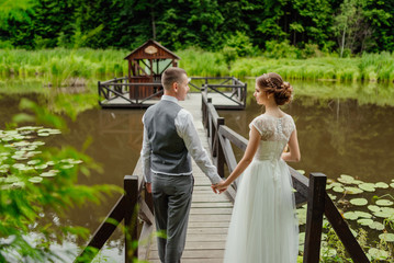 Young couple in love, groom and bride in wedding dress gainst a wooden verandah on the lake. Newlyweds hold hands and look at each other. Wedding. Wedding day. Bridal bouquet.