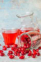      Ripe red dogwood fruits in a glass jar, next to a mug and jug with compote on an old wooden table. The concept of a healthy diet.