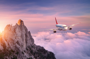 Fototapeta na wymiar Airplane is flying over clouds at sunset. Landscape with white passenger airplane, rocks, sea and purple sky with sun in summer. Passenger aircraft is landing. Business travel. Commercial plane