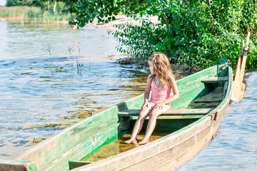 Kid girl in swimsuit sits in a boat near the shore of lake