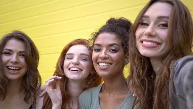 Four multiethnic beautiful female friends making selfie and showing different emotions near yellow wall