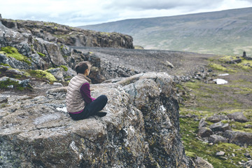 Girl in warm clothing sits on the cliff on background of mountains of Iceland.