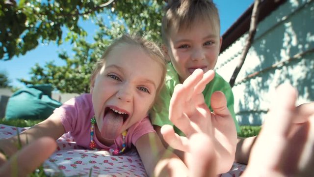 Cheerful girl and boy lying on grass in backyard and grimacing at camera showing tongues.