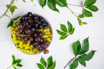 Top view bowl of various grapes: red, white and black berries and green leaves on the white wooden background. Selective focus. Space for text
