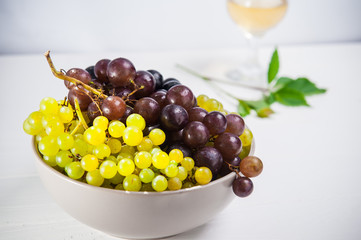 Bowl of various grapes: red, white and black berries on the white wooden table with glass of white wine and green leaf in the background . Selective focus. Space for text