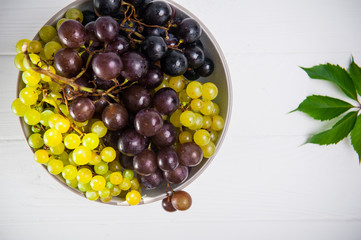 Top view bowl of various grapes: red, white and black berries and green leaf on the white wooden background. Selective focus. Space for text