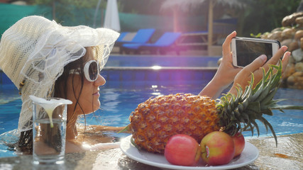 Young pretty woman wears sunglasses and hat takes selfie with mobile phone in swimming pool next to...