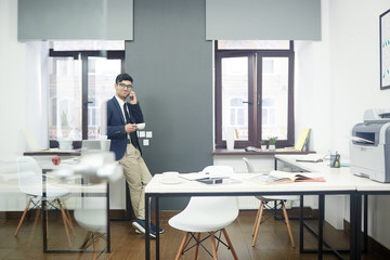 Cheerful young manager enjoying fragrant coffee and talking to his friend on smartphone while taking short break from work, interior of modern open plan office on background