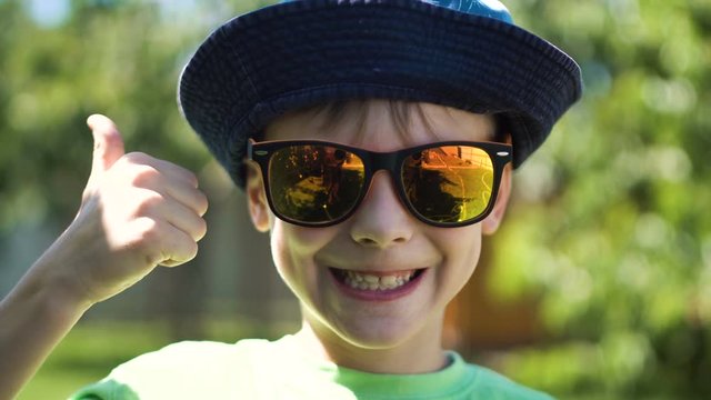 Little boy putting on colorful sunglasses and smiling at camera showing thumb up in backyard.