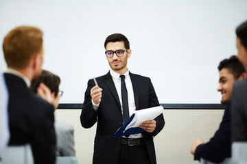 Handsome bearded speaker with clipboard in hand communicating with audience while making presentation at business conference