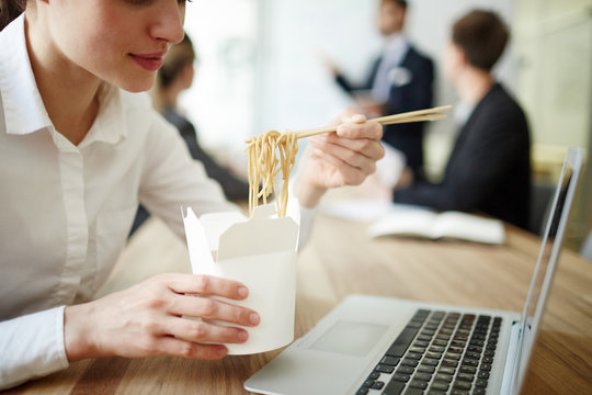 Businesswoman Eating Takeout Noodles From Box By Workplace In Front Of Laptop