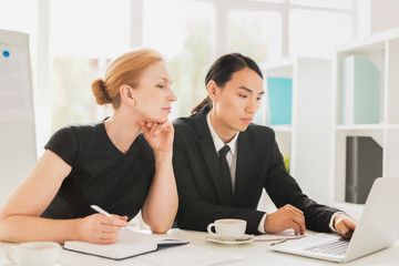 Talented young manager sharing ideas concerning promising project with his pretty superior, she listening to him with interest, interior of modern boardroom on background