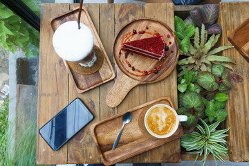 slice of red velvet cake on round wooden plate and sweet potato latte coffee and latte coffee in white cup on wooden plate with smart phone and small mock up plant.
