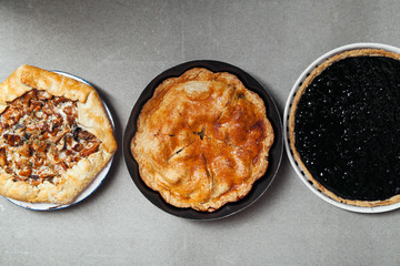 Variety of autumn pies: blueberry tart, chantarelle galette and classical apple pie with sweet pastry crust. Harvest time baking concept. Concrete rustic background. Space for text.