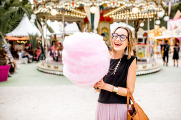 Young woman standing with pink cotton candy outdoors in front of the carrousel at the amusement park