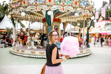 Fototapeta na wymiar Young woman standing with pink cotton candy outdoors in front of the carrousel at the amusement park