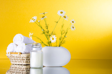 Chamomile flowers stand in a white vase.Three white towels rolled into a roll lie in a basket. Jar with cream. The background is yellow. Still life
