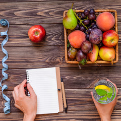 Female hands make notes in diet book on the wood table with Basket of different Fresh fruits, measuring tape, glass of water. Healthy lifestyle concept. Detox, diet. Selective focus. Copy space