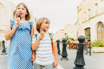 Two funny kids tourist on the street of old european city, children enjoying summer vacation on south of France. Little boy and girl eating chocolate ice cream, wearing backpacks