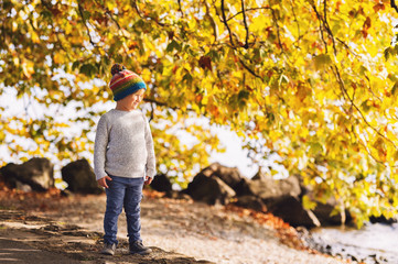 Fototapeta premium Little boy playing by the lake on a sunny day, wearing warm hat, grey knitted pullover, trousers and shoes