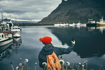 photo of girl in warm clothing with backpack on pier with background of mountains of West Fjords, Iceland. woman takes a photo on smartphone