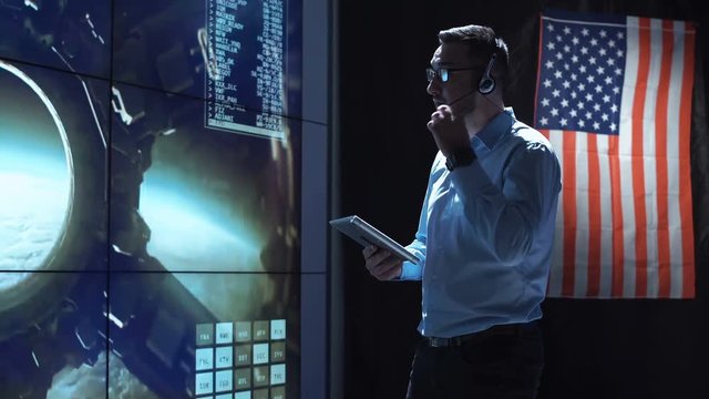 Supervisor man standing with tablet in space flight control center with American flag on background. Mars or Moon landing of spaceship. Then looking at camera. Elements of this image furnished by NASA