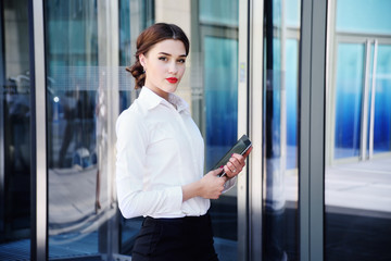 Beautiful girl in a white shirt on the background of an office building with a computer tablet in hands looking at the camera