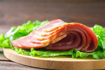 Sliced ham with fresh green lettuce leaves on a round cutting board. Meat products on a brown wooden table.