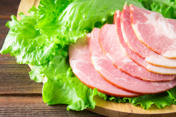 Sliced ham with fresh green lettuce leaves on a round cutting board. Meat products on a brown wooden table.