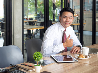 Portrait young asian businessman showing thumbs up while working with tablet, computer, documents and coffee on table at cafe office, casual business/businessman in the morning happy feeling concept