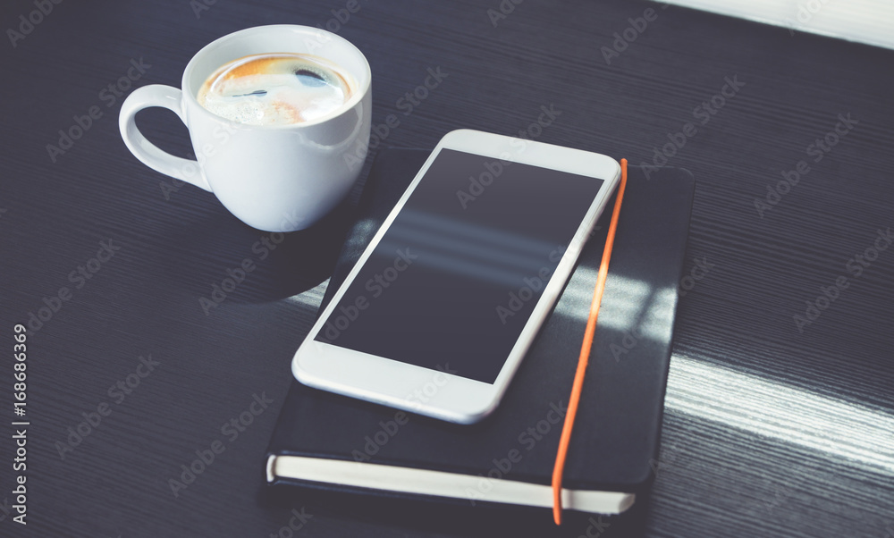 Poster blank screen of mobile phone lay on black notebook with black coffee cup near window at evening sunl