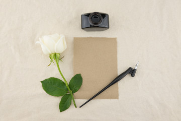 Blank brown card, oblique pen and bottle of ink decorated with white rose on white muslin fabric with copy space